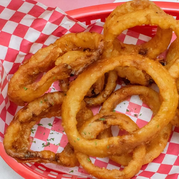 Golden-brown onion rings with parsley flakes in a red basket lined with red and white checkered paper.