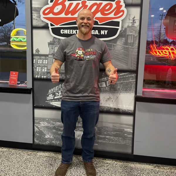 A smiling man in a "Super Fun Guy" t-shirt gives two thumbs up in front of a Rock Burger sign.