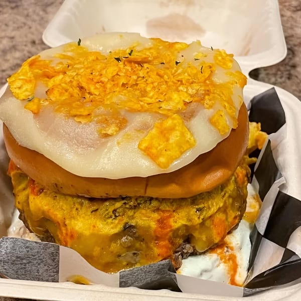 A Rock Burger with melted white cheese, crushed chips, and a thick patty in a takeout container.