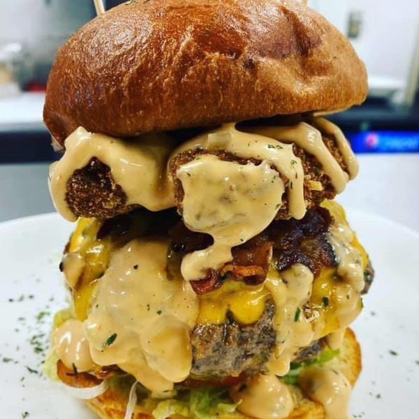 A towering Rock Burger with multiple patties, melted cheese, bacon, and a creamy sauce on a white plate.