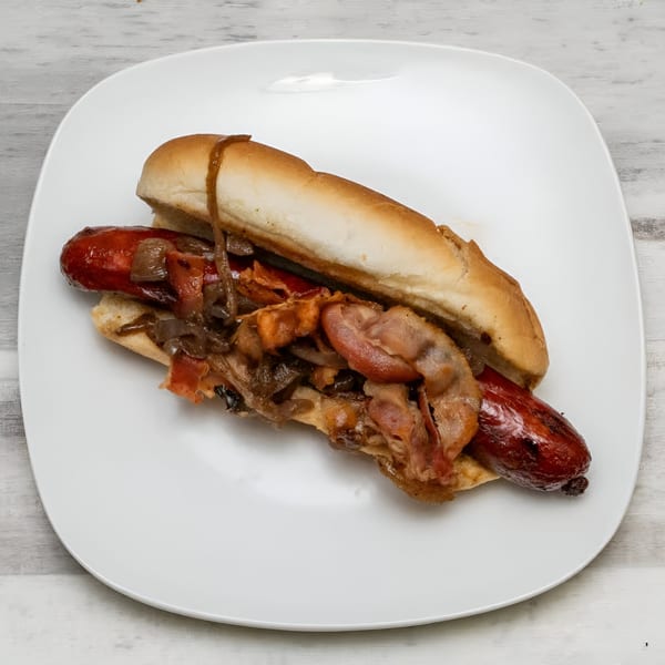 A Rock Burger hot dog with bacon, caramelized onions, and sauce on a white plate.