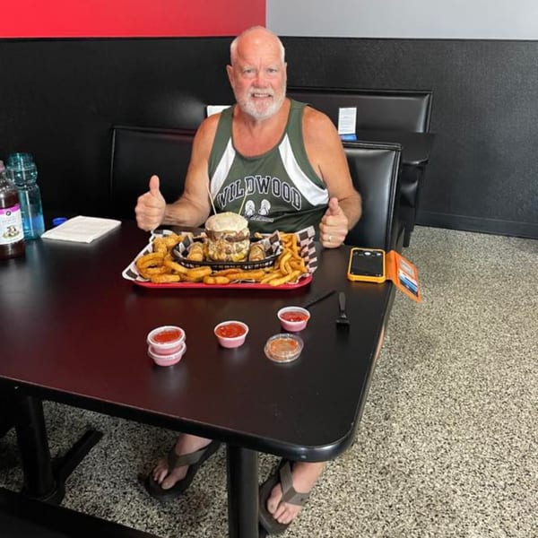 Man giving thumbs up behind a large Rock Burger meal with fries and onion rings.