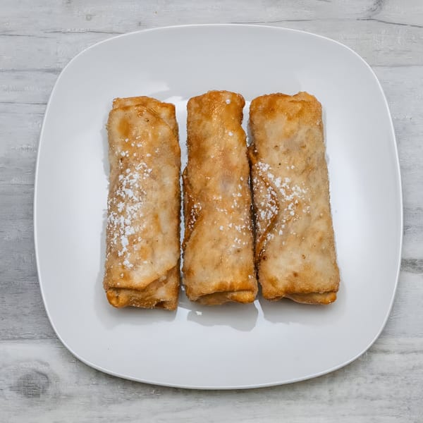 Three golden-brown fried spring rolls, lightly dusted with powdered sugar, on a white rectangular plate.