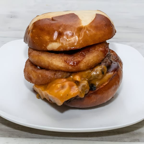 A Rock Burger with a pretzel bun, onion rings, melted cheese, and a beef patty on a white plate.