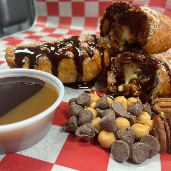 Fried dessert rolls drizzled with chocolate sauce, served with a dipping cup, chocolate chips, and pecans.