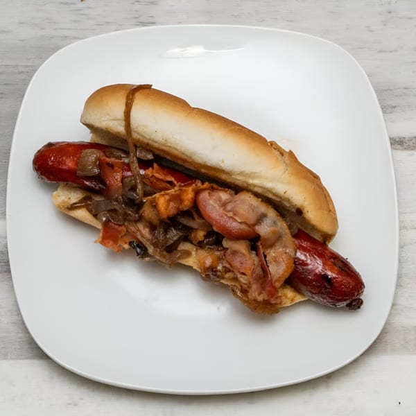Grilled hot dog with caramelized onions and bacon on a bun, served on a white plate.