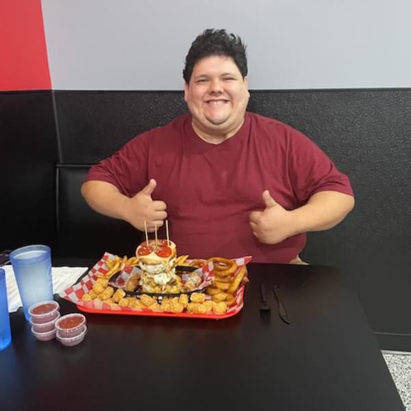 A smiling man gives two thumbs up behind a massive Rock Burger meal with fries and onion rings.